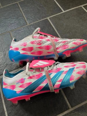 Adidas Predator rosa/blå fotbollsskor - Säljer ett par Adidas Predator fotbollsskor i vitt med rosa och blå detaljer. Skorna har en modern design med mönstrade inslag, snörning och klassisk plös med Adidas-logga. Perfekta för dig som vill sticka ut på planen och få bra grepp på konstgräs eller naturgräs.