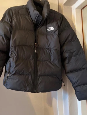 Svart pufferjacka från The North Face - Säljer en svart pufferjacka från The North Face med hög krage och dragkedja framtill. Jackan har en boxig passform och två sidofickor med dragkedja. Perfekt för kalla vinterdagar och riktigt trendig streetstil.
