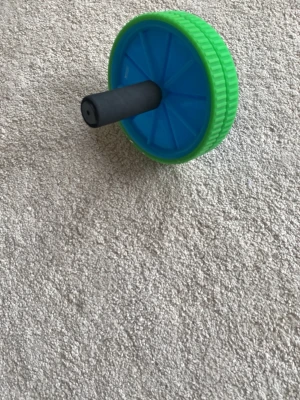 Grön och blå maghjul träningsredskap - Säljer ett maghjul med dubbla hjul i grön och blå plast samt svarta skumhandtag. Perfekt för coreträning och hemmaträning. Lätt att ta med och använda på olika underlag.