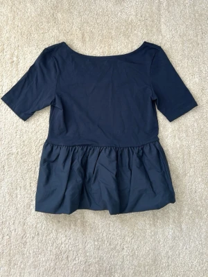 Mörkblå topp med volang - Snygg mörkblå topp med volang nedtill och halvlånga ärmar. Toppen har en enkel, rund halsringning i ryggen, men man kan ha den bak och fram. Och den är tillverkad i mjukt material som ger en avslappnad men ändå stilren look.