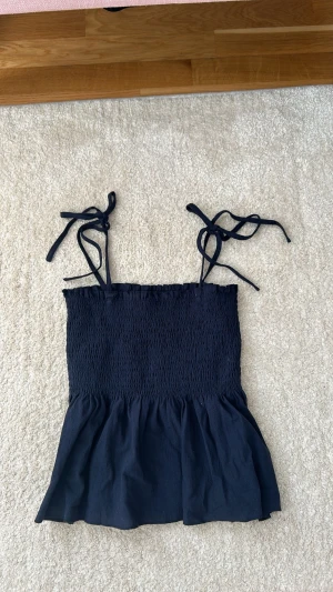 Mörkblå smockad linnetopp med knyt - Supersöt mörkblå linnetopp med smockad överdel och volang nedtill. Axelbanden knyts på axlarna för en personlig touch. Perfekt till sommaren och lätt att matcha med jeans eller kjol.