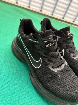 Nike Air Zoom svarta sneakers - Svarta Nike Air Zoom sneakers med vit swoosh-logga på sidan och textdetaljer. Ovandel i mesh för bra andningsförmåga och en tjock, dämpad sula för extra komfort. Klassisk snörning och sportig look som passar till streetwear.