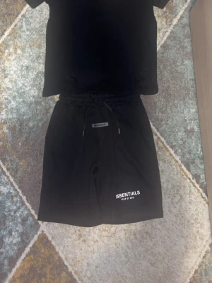 Svarta Essentials shorts med snörning - Svarta shorts från Fear of God Essentials med vit logga på vänster ben. Modellen har elastisk midja med snörning och sidofickor. Perfekta för en avslappnad streetwear-look och tillverkade i mjuk bomull.