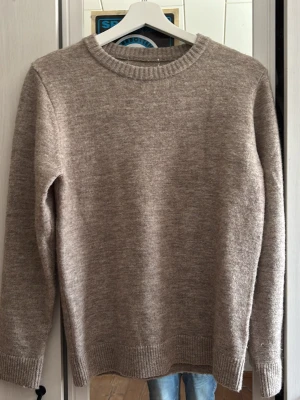 Beige stickad tröja merinoull - Mysig beige stickad tröja med rund halsringning och ribbade muddar vid ärmslut och nederkant. Tröjan har en enkel och stilren design som passar perfekt till jeans. Materialet känns mjukt och värmande, perfekt för kyligare dagar.