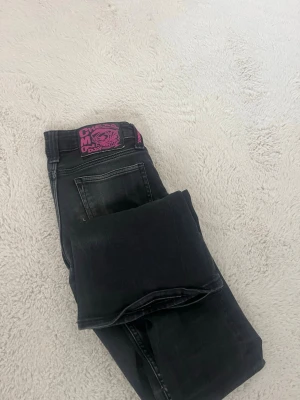 Svarta jeans från Cheap Monday - Säljer ett par svarta jeans från Cheap Monday med en cool lila patch bak. Jeansen har klassisk femficksmodell och raka ben. Materialet är denim med en lätt tvättad look. Perfekta för dig som gillar streetstyle och vill ha något som sticker ut. De är använda och har tappat lite färg, är 165 och de är lite lite långa 