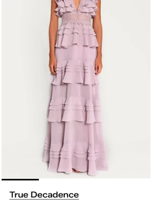 Ljuslila volangklänning True Decadence - Långklänning i ljuslila från True Decadence med flera lager volanger, djup v-ringning och spetsdetalj i midjan. Använd 1 gång! Nypris 2300kr