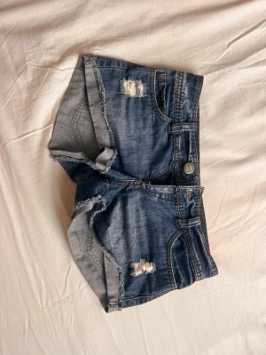Blå denimshorts med slitningar - Säljer ett par blå denimshorts med råa kanter och slitna detaljer framtill och baktill. Klassisk femficksmodell med knapp och dragkedja. Perfekta för sommaren och ger en avslappnad vibe.