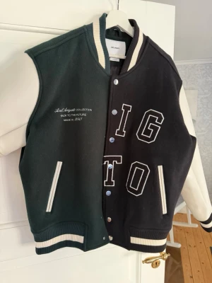 Svartgrön bomberjacka Axel arigato  - Säljer en unik svart och mörkgrön bomberjacka från axel arigato med vita kontrastärmar och broderade bokstäver fram och bak. Jackan har ribbade muddar, tryckknappar och cool retro college-stil. Materialet är en mix av tyg och konstläder på ärmarna.