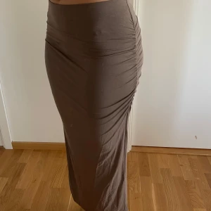 Kjol med rynkade detaljer -  En brun långkjol med hög midja och snygga rynkade detaljer längs sidan. Kjolen har en tight passform och är tillverkad i ett mjukt, stretchigt material som sitter skönt på kroppen. Perfekt att styla med en crop top eller enkel t-shirt.  Längd: 98cm