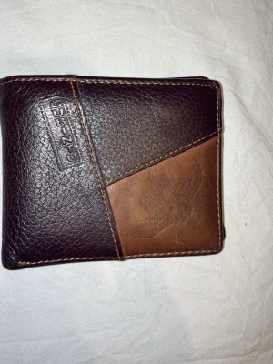 Brun plånbok i läder från Gubintu - Snygg brun plånbok i äkta läder från Subinu. Plånboken har två olika nyanser av brunt, dekorativ söm och ett präglat motiv på framsidan. Insidan har flera kortfack och ett genomskinligt fack för ID eller körkort.