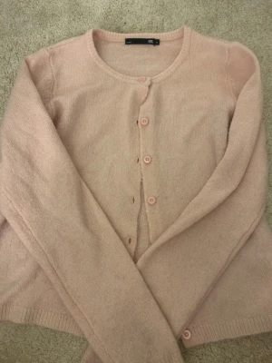 Ljusrosa kofta med rund hals - Söt ljusrosa kofta från lager 157 med rund halsringning och knappar framtill. Koftan är stickad i ett mjukt material och har långa ärmar samt ribbade muddar vid ärmslut och nederkant. Perfekt för lager på lager och enkel att matcha. Färgen ser lite annorlunda ut på bilden men annars helt nytt skick använd ett fåtal gånger. 