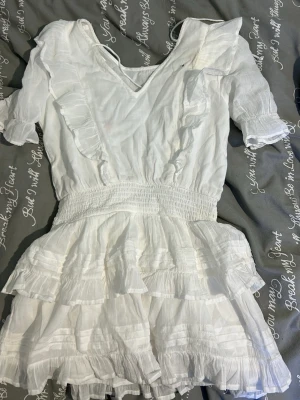 Vit volangklänning med kort ärm - Supersöt vit kortklänning med volanger och smock i midjan. Klänningen har v-ringning, korta ärmar med volangdetaljer och dubbla volanglager på kjolen. Perfekt för sommarens alla tillfällen!