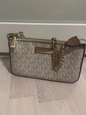 Beige kuvertväska Michael Kors - Snygg beige kuvertväska från Michael Kors med klassiskt MK-mönster, guldfärgad kedja och dragkedja. Väskan har en metallbricka med logga framtill och detaljer i brunt läder. Perfekt storlek för det viktigaste och ger en lyxig känsla.