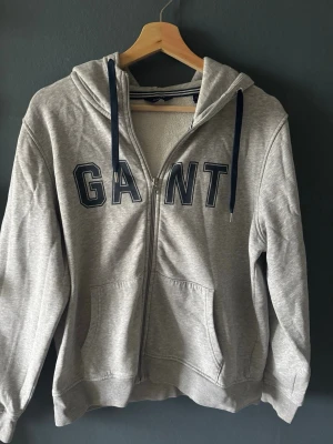 Grå zip hoodie från GANT - Snygg grå hoodie från GANT med dragkedja och stora blå bokstäver på bröstet. Mjuk insida i fleece, två fickor fram och justerbar huva med mörkblå snören. Har två fläck på framficka, men det inget man märker🩷. Perfekt för en avslappnad och sportig stil.
