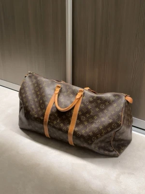 Louis Vuitton Keepall 60 Monogram - En Louis Vuitton Pre-Owned Keepall Bandoulière 60 Monogram i gott skick. En snygg väska i brun färg som är perfekt för resor. En sällsynt modell som är svår att få tag på. Det finns några mindre defekter (se bilder), såsom repor på väskans läderrem, men inga hål eller liknande skador. Fortfarande i mycket gott skick.  100% automatisk. Bevis finns självklart. Sedan serienummer på väskan osv…  Vid fler frågor är det bara att höra av er! MVH 