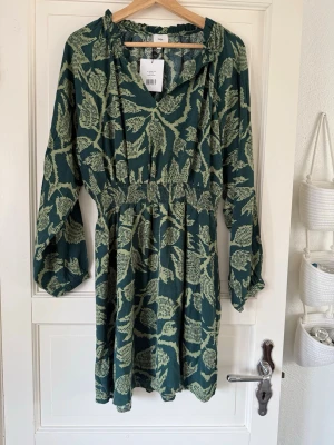  klänning från Ange - Grön kortklänning från Ange med ljusgrönt blommigt mönster. Klänningen har v-ringning, långa ärmar och resår i midjan för en snygg passform. Tillverkad i ett mjukt och lätt material som känns skönt mot huden.