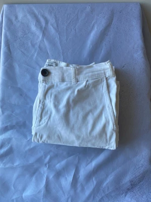 Vita chinos från Les Deux - Snygga vita chinos från Les Deux med klassisk passform och elastisk midja. Byxorna har sidofickor och en enkel knappstängning framtill. Perfekta för en clean och stilren look. Materialet känns mjukt och bekvämt. Stora i stl passar en man i M!