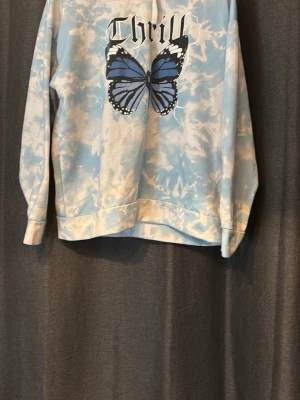 Blå tie-dye hoodie med fjärilstryck - Säljer en blå och vit tie-dye hoodie med stort tryck av en blå fjäril och texten 'Thrill' på bröstet. Hoodien har långa ärmar och en avslappnad passform. Perfekt för dig som gillar unika och trendiga plagg med coolt motiv.