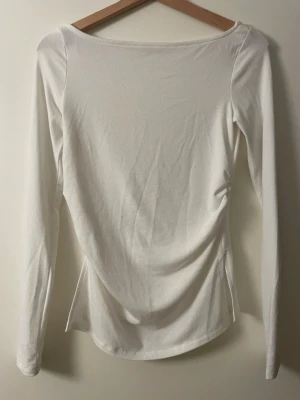 Berhska Vit långärmad topp med rynkdetalj - Säljer en enkel och stilren vit långärmad topp med rynkdetalj på sidan. Toppen har en rundad halsringning och är gjord i mjukt bomullsmaterial som känns skönt mot huden. Perfekt att matcha med jeans eller kjol för en clean look.