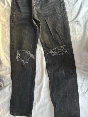 Svarta slitna jeans med hål - Säljer ett par svarta jeans med raka ben och stora slitna hål på båda knäna. Jeansen har en cool, avslappnad vibe och är perfekta för dig som gillar streetstyle. Materialet är klassisk denim och färgen är tvättad svart.