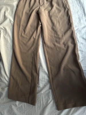 Beiga vida kostymbyxor - Säljer ett par beiga kostymbyxor med rak och vid passform. Byxorna har hög midja och är tillverkade i ett mjukt, vävt material som ger en snygg siluett. Perfekta för en clean och stilren look.