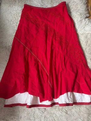 Röd långkjol med broderade detaljer - Snygg röd långkjol med vita broderade blommor och dekorativa sömmar. Kjolen har en vit underkjol som sticker fram nedtill och vågig kant. Perfekt för dig som vill ha en färgstark och unik look.