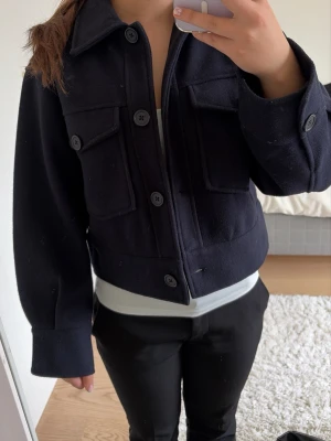 Mörkblå ull-overshirt/kappa med fickor - Stilren mörkblå overshirt/kappa i ullmix med stora knappar framtill och två rejäla bröstfickor med lock. Kortare modell med klassisk krage och raka ärmar. Perfekt lager på lager-plagg för en trendig look.