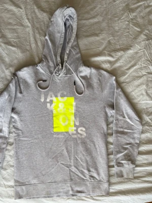 Jack & Jones hoodie  - Säljer en stilren grå hoodie från Jack and Jones med ett modernt tryck i vitt och neon på bröstet. Modellen heter Jconep Sweat Hood och är i herrstorlek M. Tröjan har en mjuk fleece insida, justerbar luva med band och ribbade muddar. Materialet är 93% bomull och 7% viskos vilket ger en skön kvalitet. Den är i mycket gott skick, använd ca 3 gånger. 