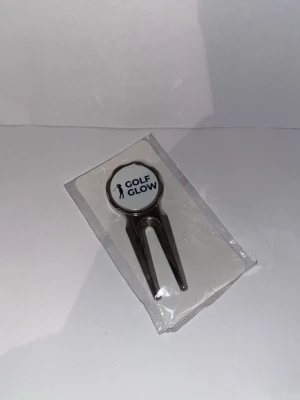 Golf Glow greenlagare  - Säljer ett markeringsverktyg från Golf Glow i metall med två piggar och rund topp. Verktyget har en vit och blå logga på ovansidan och är perfekt för golfspelare som vill fixa nedslagsmärken på greenen.