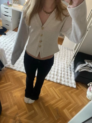 Beige kofta med guldfärgade knappar - Snygg beige kofta med v-ringning och fyra stora guldfärgade knappar framtill. Modellen är något croppad och har långa ärmar. Perfekt att styla med jeans eller kjol för en trendig look.