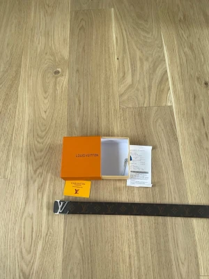 Hm brunt  skärp - Säljer ett exklusivt hm skärp med klassiskt brunt monogrammönster och silverfärgat hm-spänne. Skärpet kommer med originalbox och kvitto. Perfekt för dig som vill ha en ikonisk accessoar med lyxig känsla.