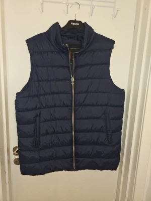 Mörkblå dunväst med dragkedja - Snygg mörkblå dunväst med hög krage och horisontella sömmar. Västen har två fickor med dragkedja och stängs med en silverfärgad dragkedja framtill. Perfekt för lager-på-lager och ger en clean look.