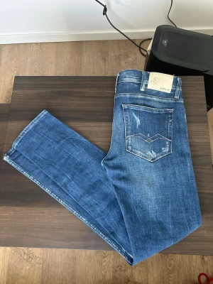 Blå slitna jeans från Replay - Säljer ett par blå jeans från Replay med slitningar och distressed detaljer både fram och bak. W31 L32 Klassisk femficksmodell med raka ben och normal midja. Snygga kontrastsömmar och Replay-lapp bak i midjan. Perfekta för en avslappnad och trendig look.