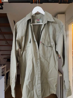 Beige skjorta från Levi's - Klassisk beige skjorta från Levi's i 100% bomull. Skjortan har lång ärm, bröstficka med knapp och den ikoniska röda Levi's-taggen. Perfekt passform och tidlös design med tydlig vintagekänsla. Pris kan alltid diskuteras ! 