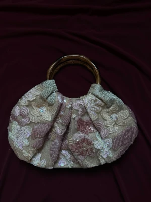 Blommig handväska med paljetter - Unik handväska med broderade blommor i beige, rosa och lila toner. Väskan har ett handtag i bambu och är dekorerad med skimrande paljetter som ger extra glans. Perfekt accessoar för dig som vill sticka ut med en romantisk och bohemisk stil.