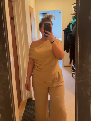 Beige off shoulder jumpsuit - Trendig beige jumpsuit med off shoulder-design och vida ben. Tillverkad i ett mjukt, lätt tyg som ger en snygg drapering över höften. Perfekt för dig som gillar en stilren och modern look. Inga synliga fickor eller dragkedjor. Passar till sneakers eller klackar.