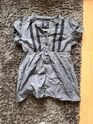 Blå kortärmad blus med knappar - Snygg grå blus från Sublevel med korta puffärmar och dekorativa svarta spetsdetaljer framtill. Blusen har knappar längs bröstet och en markerad midja för en feminin siluett. Perfekt till jeans eller kjol för en trendig look.