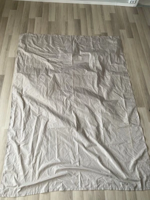 Beige duschdraperi i rektangulär form - Stort beige duschdraperi i rektangulär form. Materialet ser ut att vara tunt och lätt, troligen bomull eller polyester. Enkel design utan mönster eller tryck.