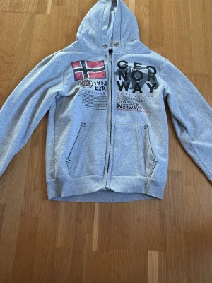 Grå hoodie från Geo Norway med tryck - Snygg grå hoodie från Geo Norway med dragkedja, huva och två fickor framtill. Stor norsk flagga och coola tryck på bröstet ger en sportig vibe. Perfekt för dig som gillar streetwear och vill sticka ut lite extra. Mjuk insida för extra komfort.