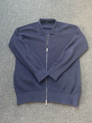 Mörkblå ziptröja från Massimo Dutti - Snygg mörkblå ziptröja från Massimo Dutti med klassisk krage och hel dragkedja framtill. Tröjan är stickad i ett mjukt material och har långa ärmar samt ribbade muddar. Perfekt för en stilren och avslappnad look.