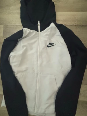 Nike svartvit hoodie med dragkedja - Nike hoodie i svart och vitt med dragkedja framtill och klassisk logga på bröstet. Huva och fickor framtill. Ärmar och huva är svarta medan kroppen är vit. Perfekt för en sportig och avslappnad stil.