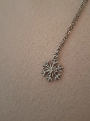 Silverfärgat halsband med snöflinga - Snyggt halsband i silverfärg med ett hänge format som en snöflinga. Hänget har små gnistrande stenar som ger extra bling och en vintrig känsla. Kedjan är tunn och elegant, perfekt för att lyfta vilken outfit som helst.