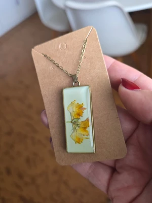 Halsband med inneslutna gul blomma - Unikt halsband med rektangulärt hänge i metall och transparent resin där en gul blomma är inneslutna. Kedjan är tunn och 50 cm längt, och hänget har en modern, minimalistisk känsla. Perfekt för dig som gillar naturinspirerade accessoarer och vill sticka ut med något personligt.