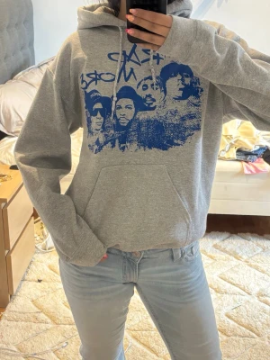 Grå hoodie med blått tryck från Gildan - Säljer en grå hoodie från Gildan med ett stort blått tryck framtill. Tillverkad i mjuk bomullsblandning, perfekt för en avslappnad streetwear-look.