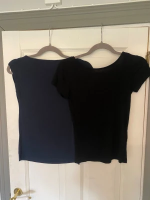 Två bas-toppar i svart och marinblå - Säljer två stilrena toppar, en i svart och en i marinblå. Den svarta toppen har korta ärmar och djup rygg, medan den marinblå är ärmlös med enkel, rak passform. Perfekta att matcha till olika outfits.