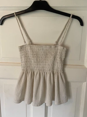 Vitt topp från Gina Tricot - Säljer en söt vit topp från Gina Tricot med smockad överdel och tunna justerbara axelband. Toppen har en luftig och flowy passform nedtill, perfekt för varma dagar. Materialet känns lätt och bekvämt mot huden.