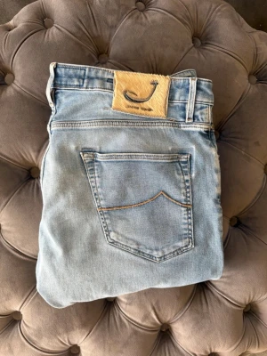 Ljusblå jeans från Jacob Cohen - Säljer ett par ljusblå jeans från Jacob Cohen med klassisk femficksmodell och knappgylf. Jeansen har en snygg tvättad look, kontrastsömmar och patch med logga bak i midjan. Perfekta för en avslappnad och trendig stil. 