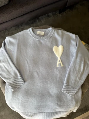 Ljusblå stickad tröja från Ami Paris - Säljer en ljusblå stickad tröja från Ami Paris med ett stort vitt hjärta och bokstaven A på bröstet. Tröjan har rund halsringning, ribbade muddar och är tillverkad i mjukt material. Perfekt för dig som gillar stilrena detaljer.