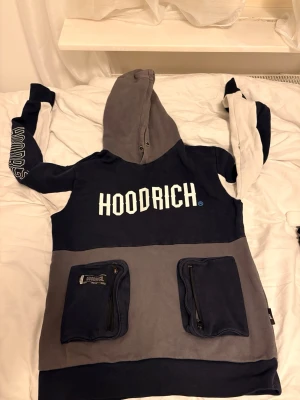 Hoodrich hoodie i blått och grått - Snygg hoodie från Hoodrich i blått och grått med stor logga på bröstet och ryggen. Två stora fickor med dragkedja framtill och tryck på ärmarna. Perfekt för en streetwear-look. Huva med dragsko och cool färgblock-design.