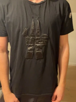 Svart Moncler t-shirt med stor logga - Snygg svart t-shirt från Moncler med stor ton-i-ton logga på bröstet och klassisk Moncler-patch på ärmen. T-shirten har rund hals och korta ärmar, tillverkad i mjuk bomull för skön känsla. Perfekt för dig som gillar stilrena plagg med exklusiv touch.